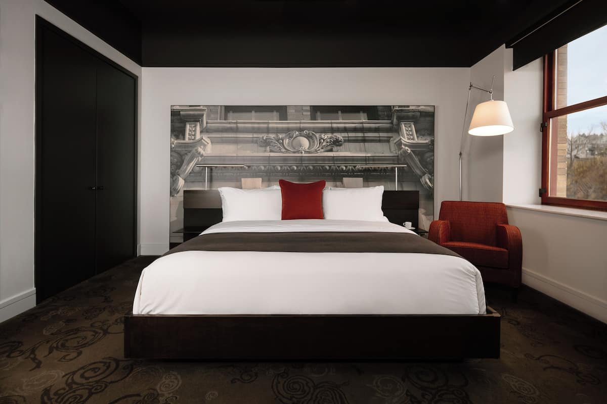 A modern hotel room with a large bed, a red chair, a decorative headboard, and a large window.
