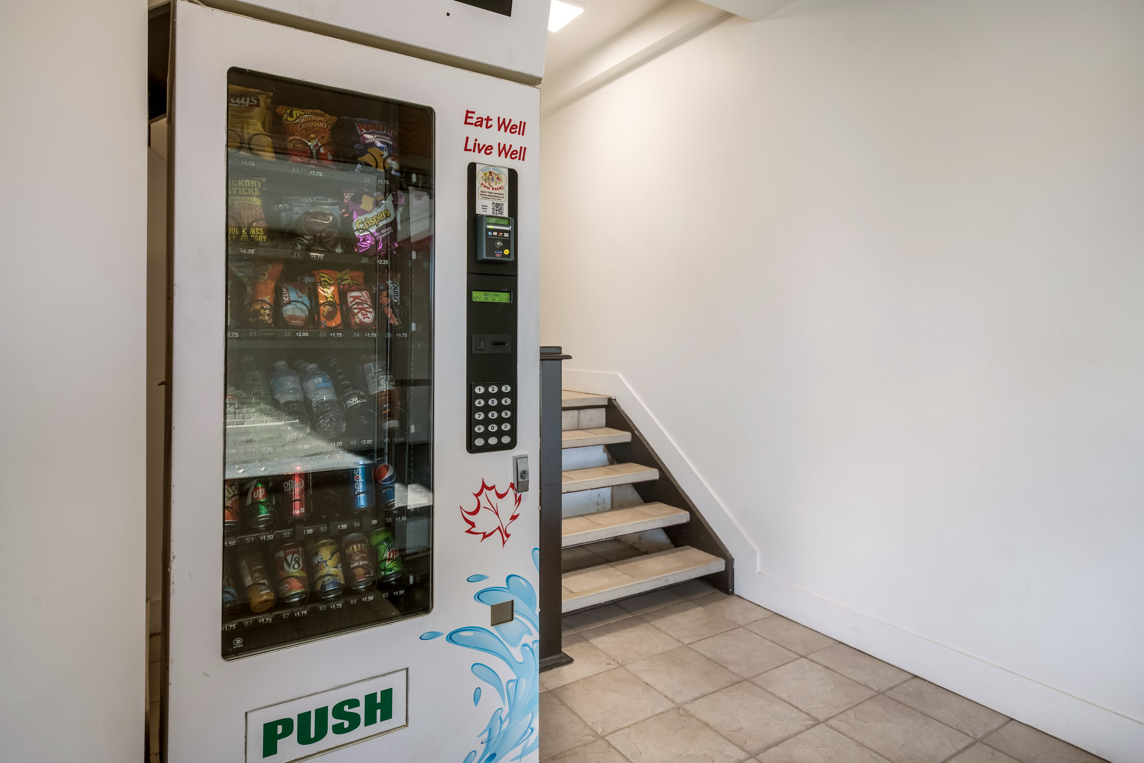 En automat med en 'PUSH' knap, placeret ved siden af et trappeopgang i et lyst, hvidt rum.