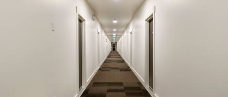A hotel corridor with white walls, carpeted flooring, and doors on both sides.