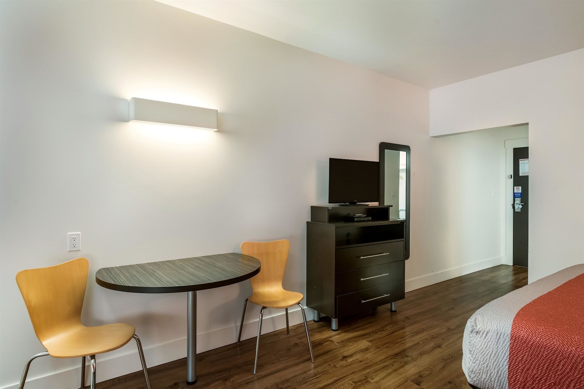 Une chambre d’hôtel avec un plancher en bois, une table ronde avec deux chaises, un meuble noir pour la télévision avec un écran plat, et un lit recouvert d’une couverture à motifs.
