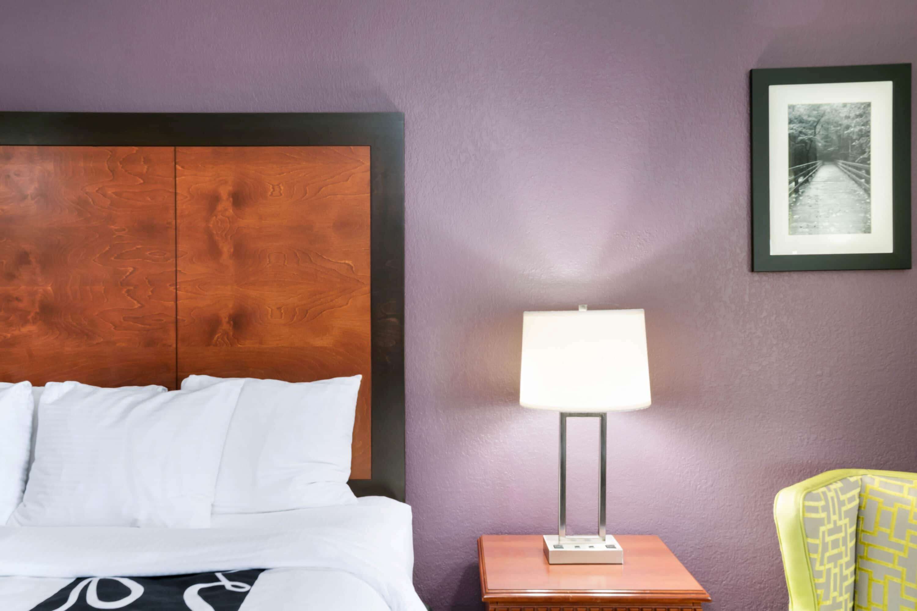 A hotel room with a wooden headboard, a bedside table with a lamp, a framed picture on the wall, and a patterned chair.