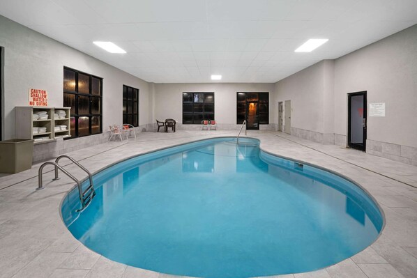 Una piscina interior con superficie de agua cristalina, rodeada por un suelo y paredes de azulejos. Hay carteles en las paredes que indican precaución y prohíben el clavado.