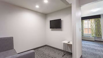A hotel room with a grey sofa, a small table, a flat-screen TV mounted on the wall, and a patterned carpet.