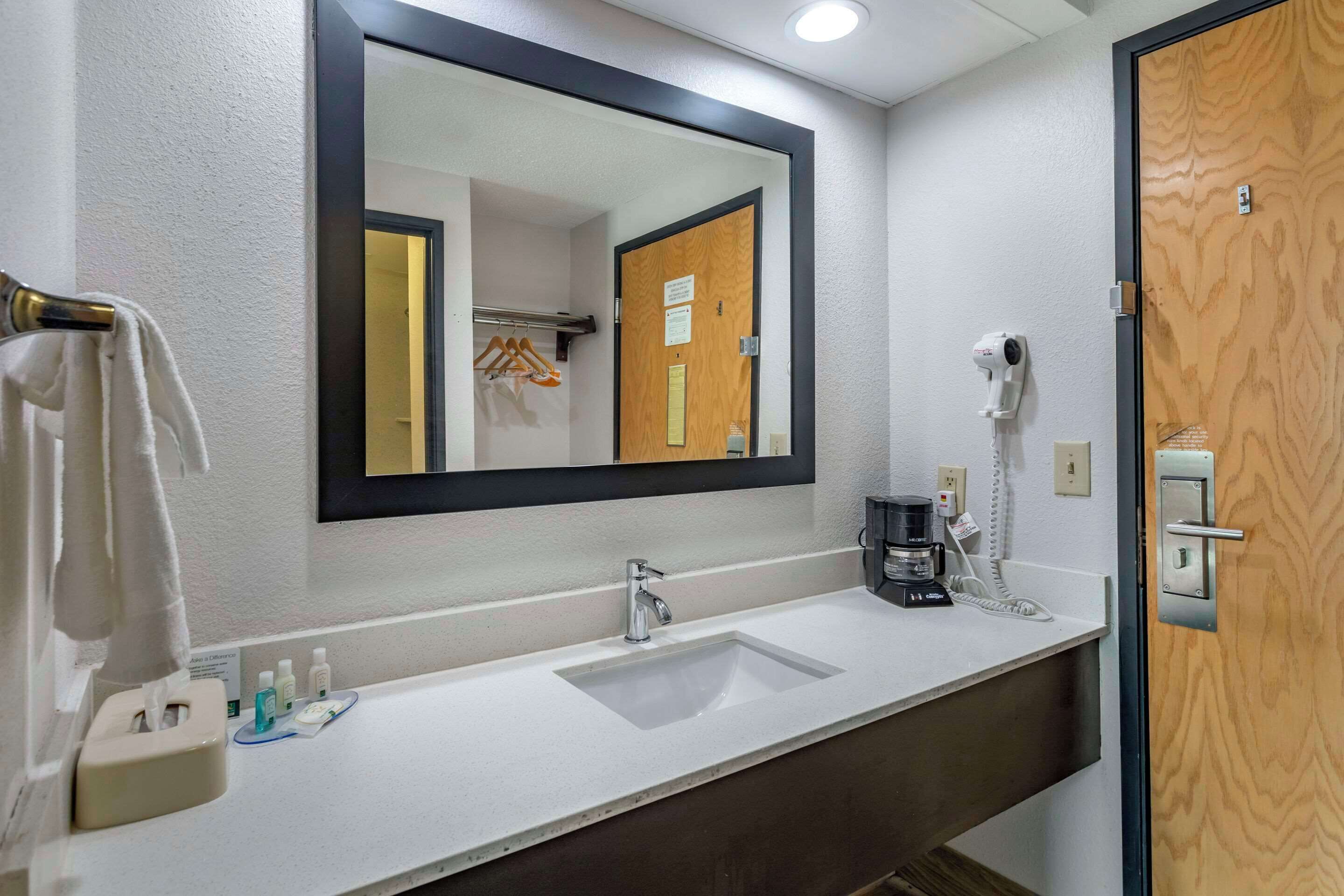 Un baño de hotel con un lavamanos blanco, un espejo, un perchero para toallas, un secador de pelo y una cafetera.