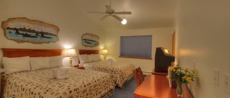 Une chambre d’hôtel avec deux lits, un bureau en bois, un ventilateur de plafond et des œuvres d’art accrochées aux murs.