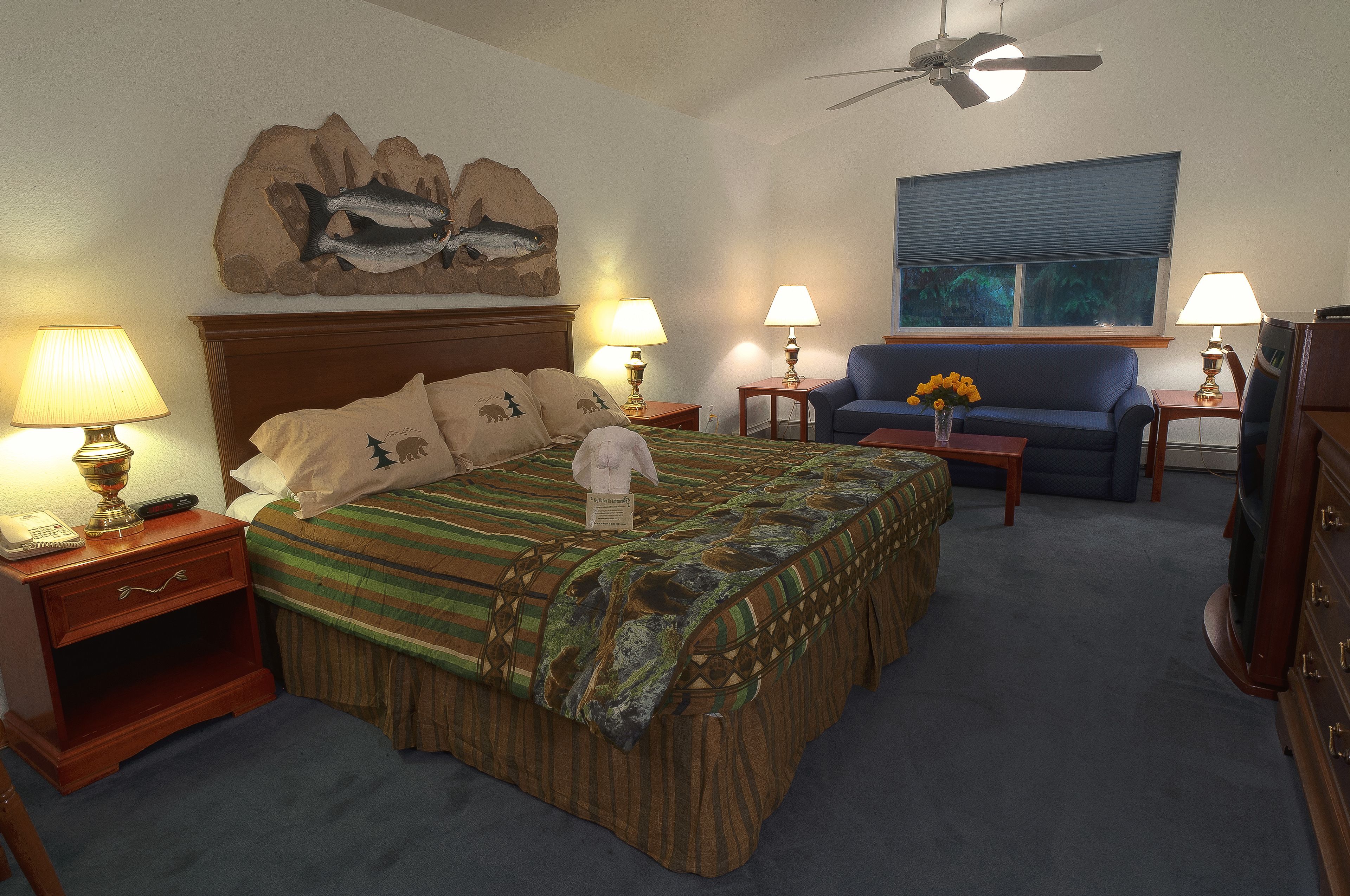 A hotel room with a bed, two bedside tables with lamps, a blue sofa, a television, and a ceiling fan.
