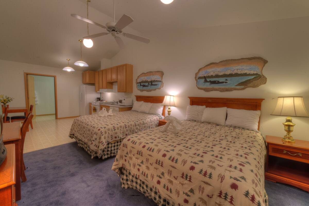 A bedroom with two beds, a ceiling fan, and a kitchenette in the background.