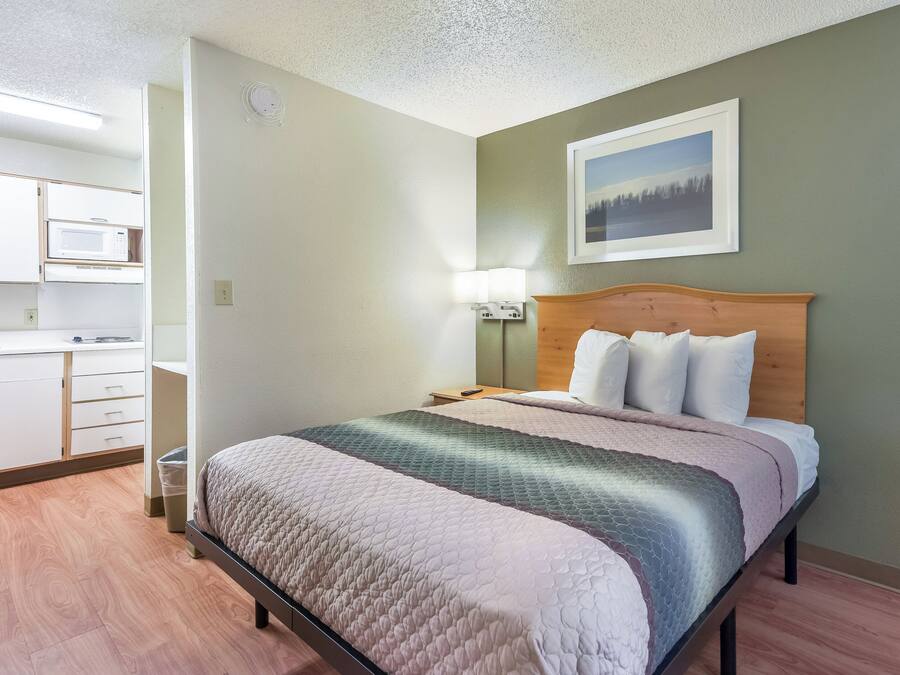 A hotel room with a bed, a nightstand, a lamp, and a view of the kitchen.