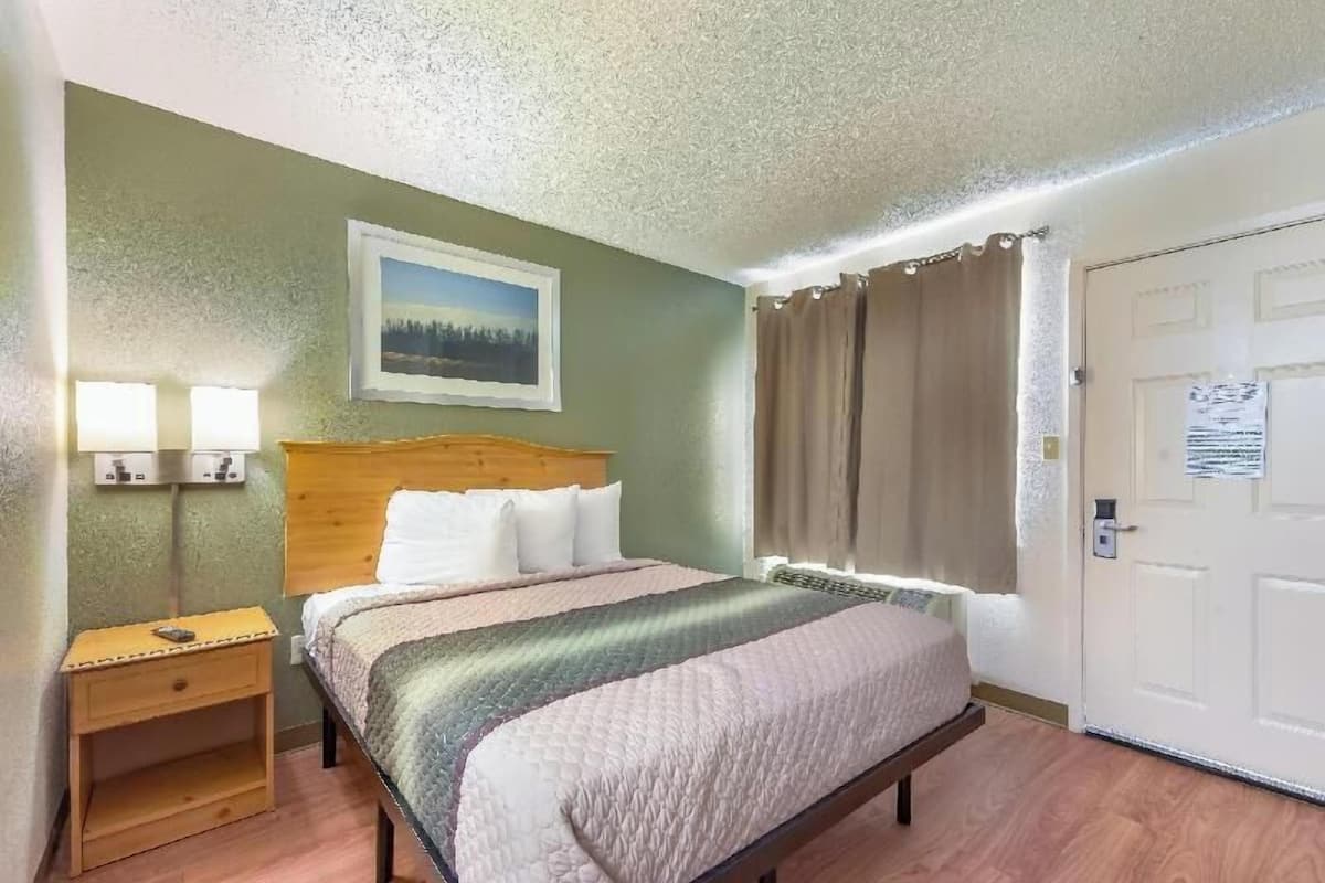 A hotel room with a bed, bedside lamps, a painting, a door, and a window with curtains.
