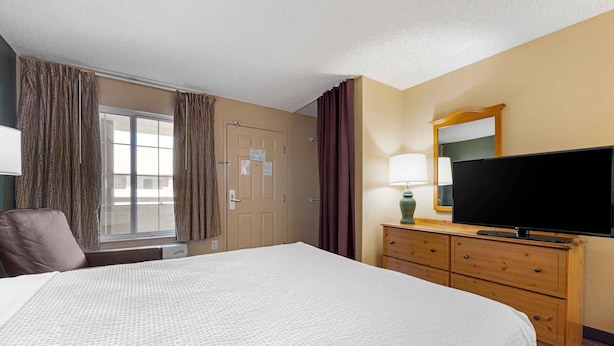 A hotel room with a large bed, a television on a wooden dresser, a window with curtains, and a door with a sign.