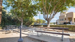 An outdoor seating area with benches and picnic tables, surrounded by trees and a building in the background.