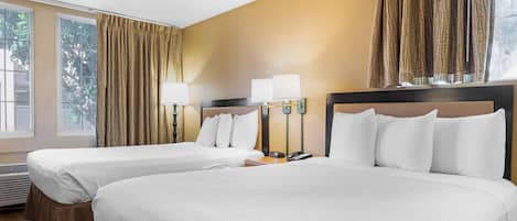 A hotel room with two beds, a window with curtains, a bedside table with lamps, and a thermostat.