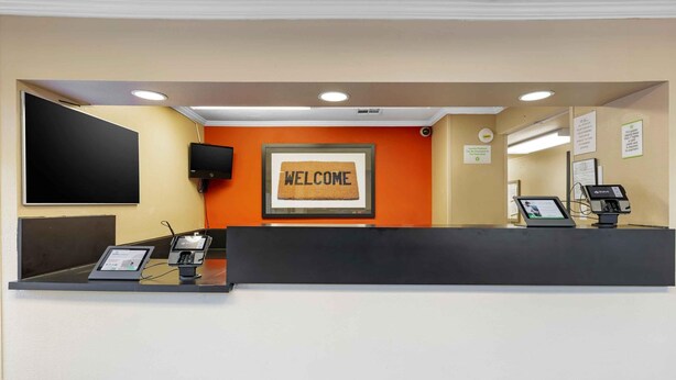 A hotel reception desk with a 'WELCOME' sign, a flat-screen TV, a computer, and a payment terminal. - Extended Stay America Suites San Diego Fashion Valley (San Diego)