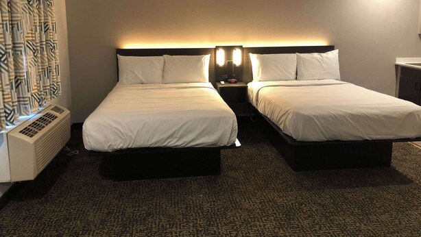 A hotel room with two beds, a nightstand with a lamp, a heater, and a patterned curtain. - Studio 6 Killeen, TX (Killeen)