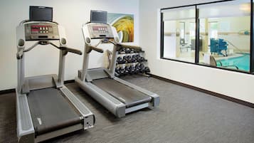 Dos cintas de correr en un gimnasio con un ventanal grande, un televisor de pared y un estante con mancuernas.
