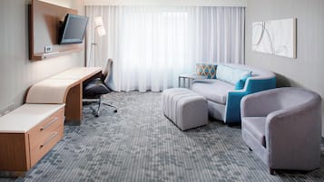 A hotel room with a desk, a sofa, an ottoman, a chair, a TV mounted on the wall, and a window with curtains.