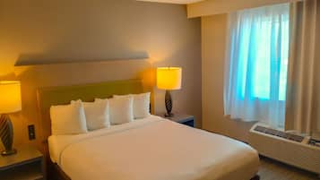 A hotel room with a large bed, two bedside lamps, a window with curtains, and a wall-mounted air conditioner.
