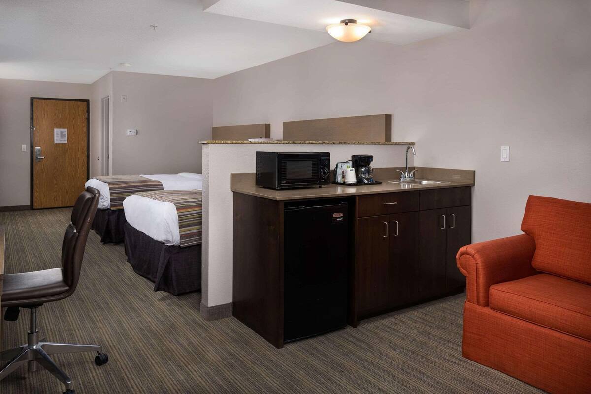A hotel room with a kitchenette, a bed with a striped bedspread, a brown chair, and a red sofa.