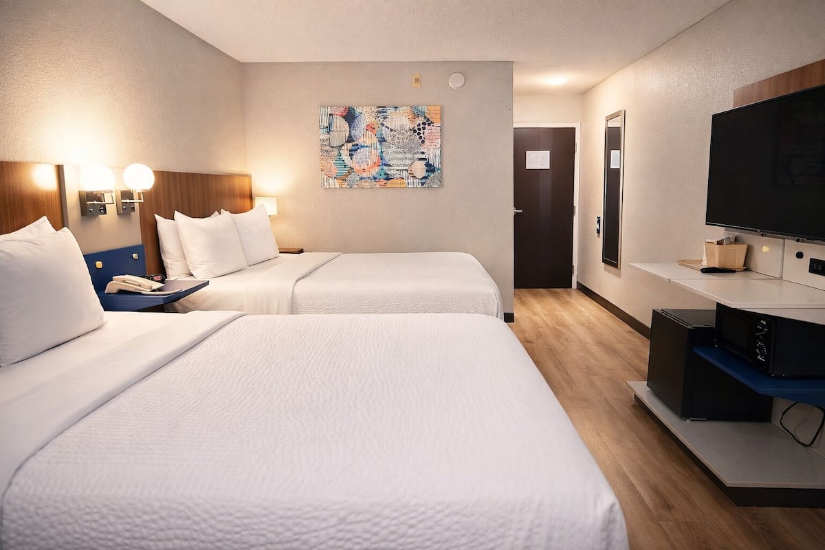 A hotel room with two beds, a desk with a flat-screen TV, a painting on the wall, and a wooden floor.