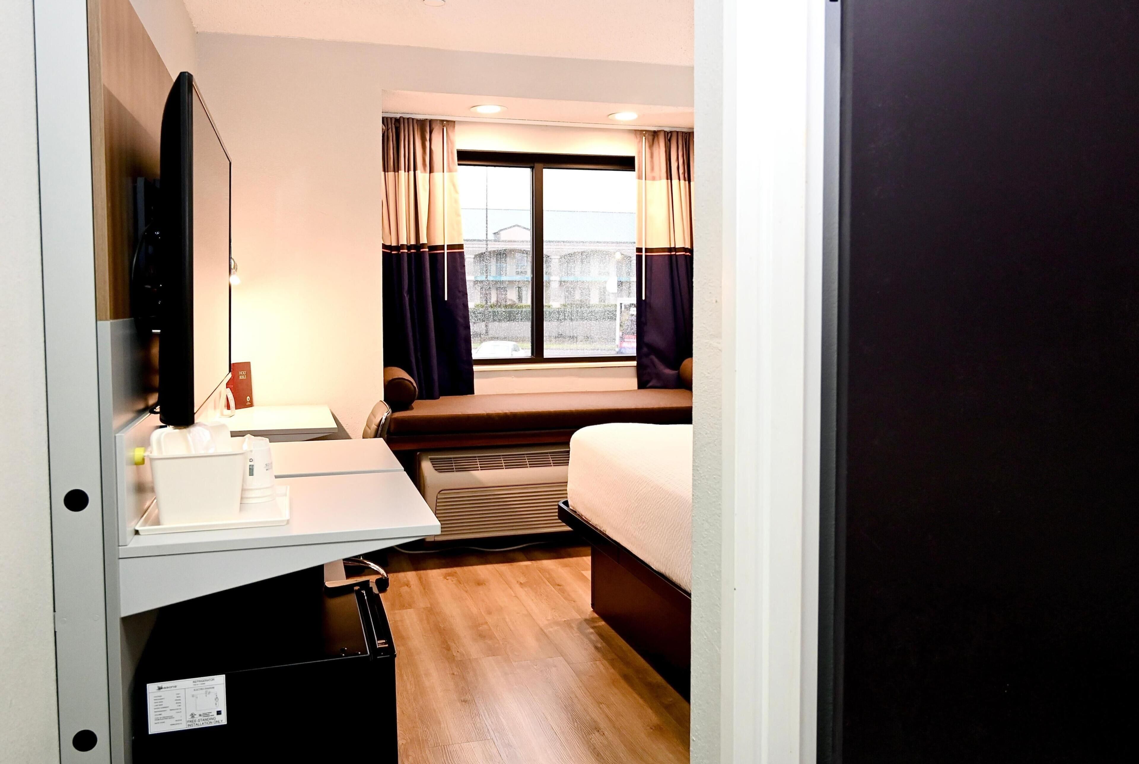 A hotel room with a bed, a desk with a computer, a TV, and a window with curtains.