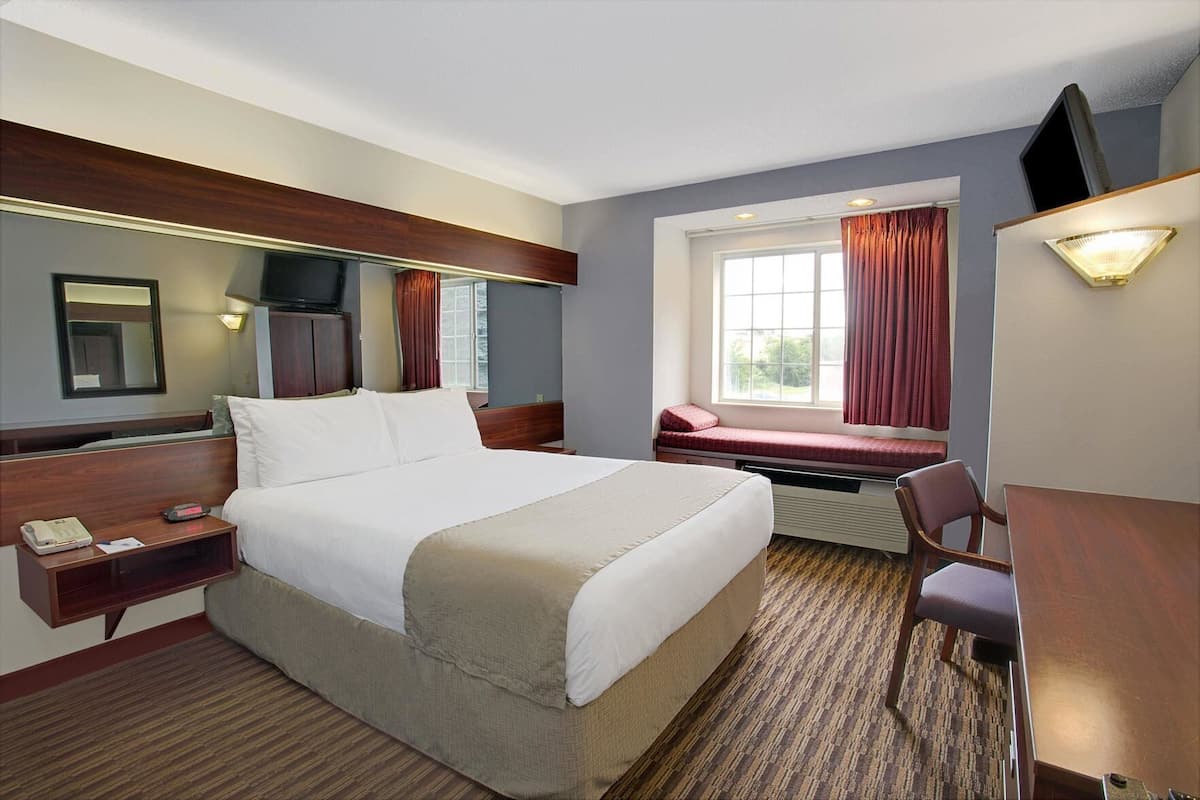 A hotel room with a bed, bedside table, desk, chair, TV, window with curtains, and a mirror.