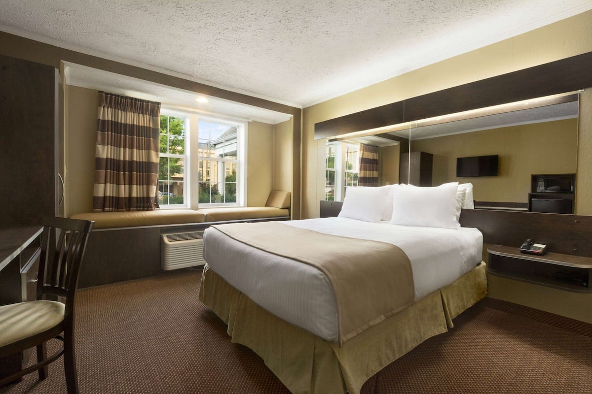 A hotel room with a bed, a desk, a chair, a window with curtains, a television, and a mirror.