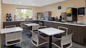 A modern kitchen area with white tables and chairs, equipped with coffee makers, a refrigerator, and a juice dispenser. - Microtel Inn & Suites by Wyndham Columbia/Fort Jackson N (Columbia)