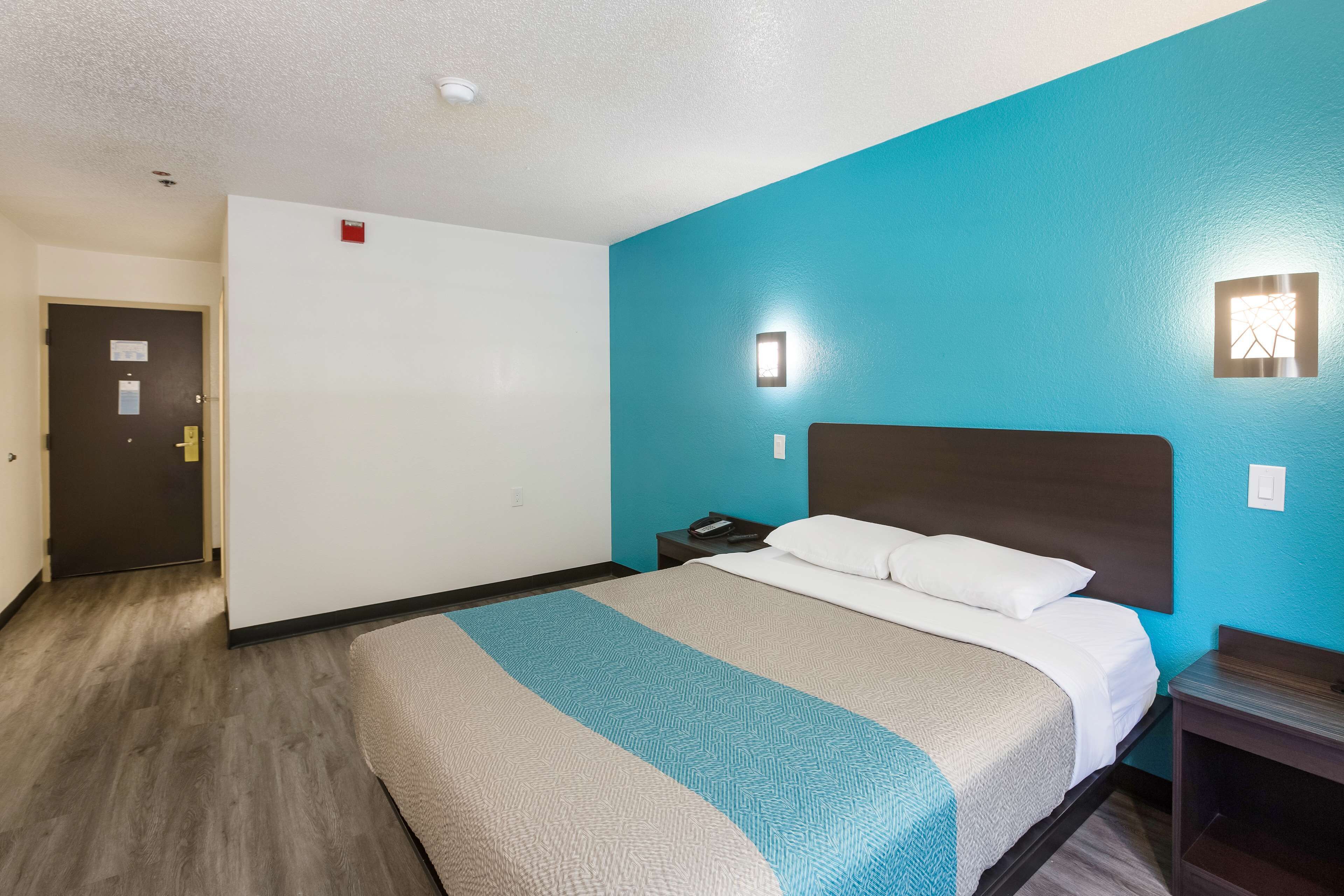 Una habitación de hotel con una cama, una mesita de noche, una lámpara fijada a la pared y una puerta.