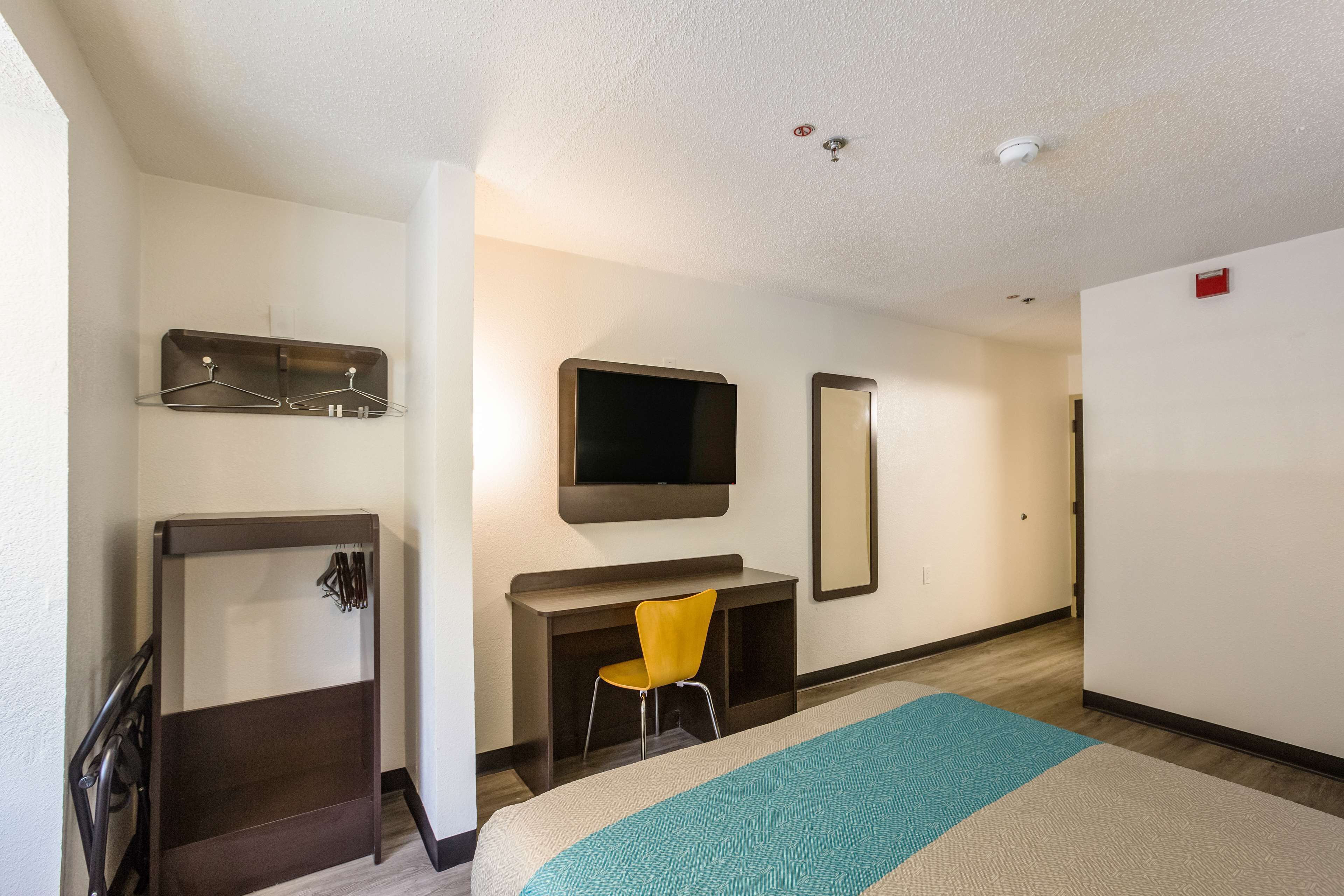 A hotel room with a bed, a desk with a chair, a TV mounted on the wall, and a coat rack.