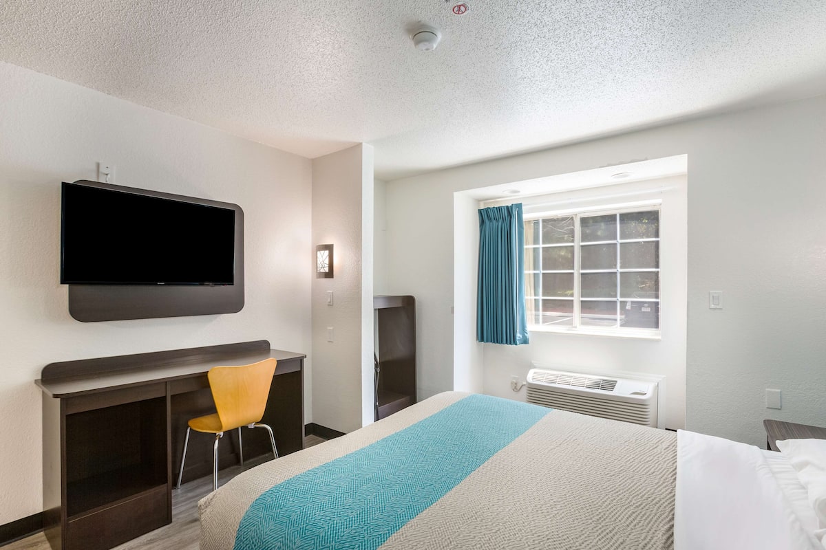 A hotel room with a bed, a TV mounted on the wall, a desk with a chair, a window with curtains, and a fireplace.