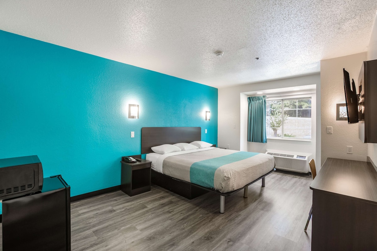 A modern hotel room with a teal accent wall, a bed with a wooden headboard, a desk, and a window with curtains.