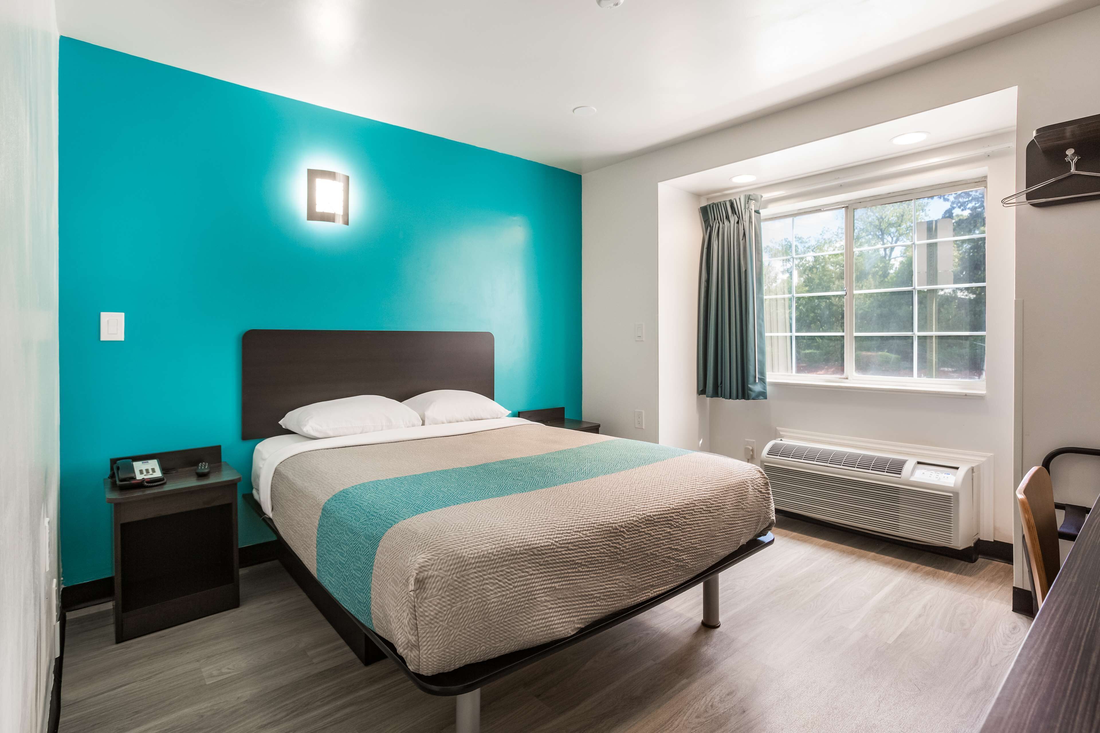 A modern hotel room with a teal accent wall, a bed with a beige and teal bedspread, a bedside table with a phone, a window with curtains, and a wall-mounted heater.