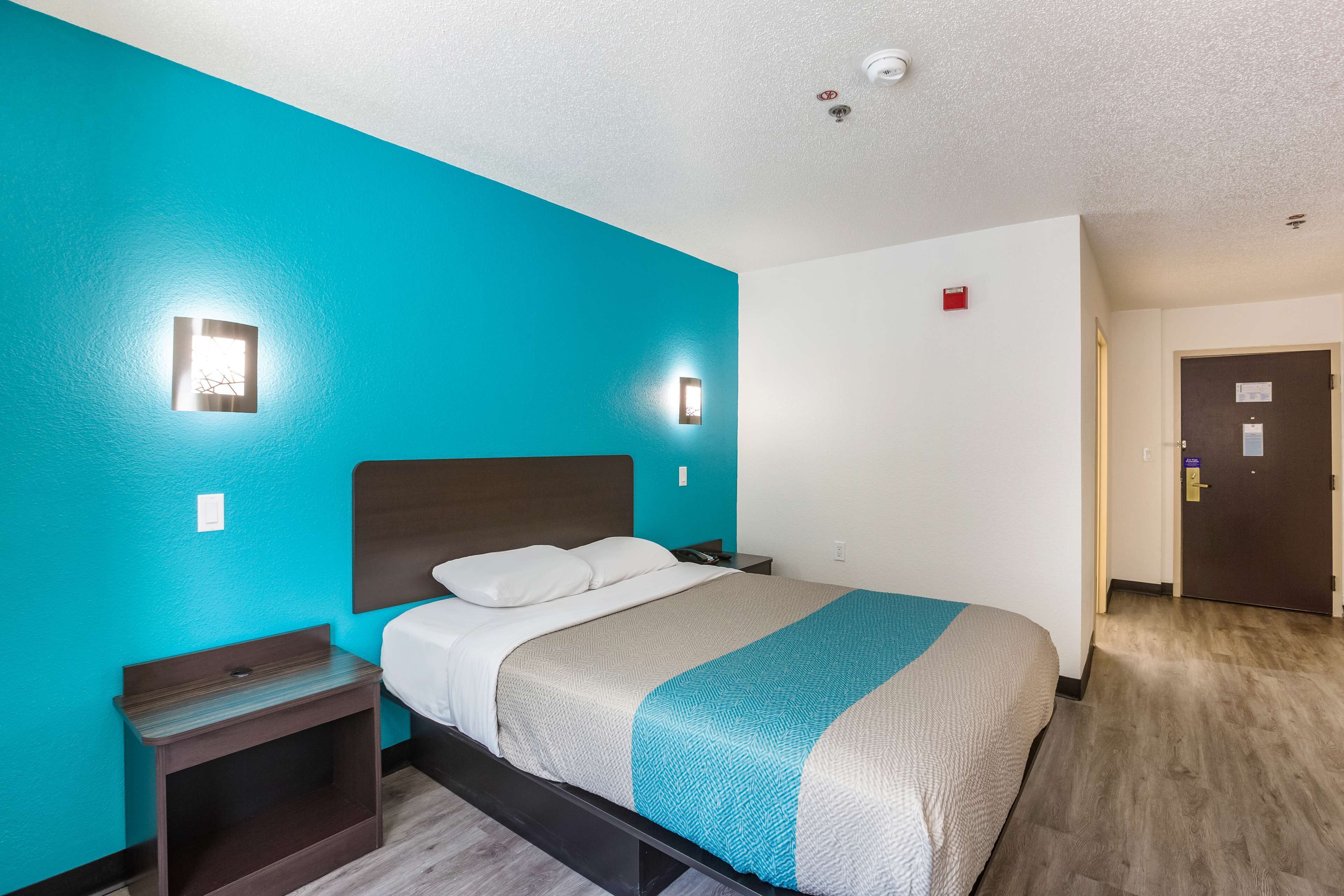 Una habitación de hotel con una cama, una mesita de noche, una lámpara de pared y una puerta.