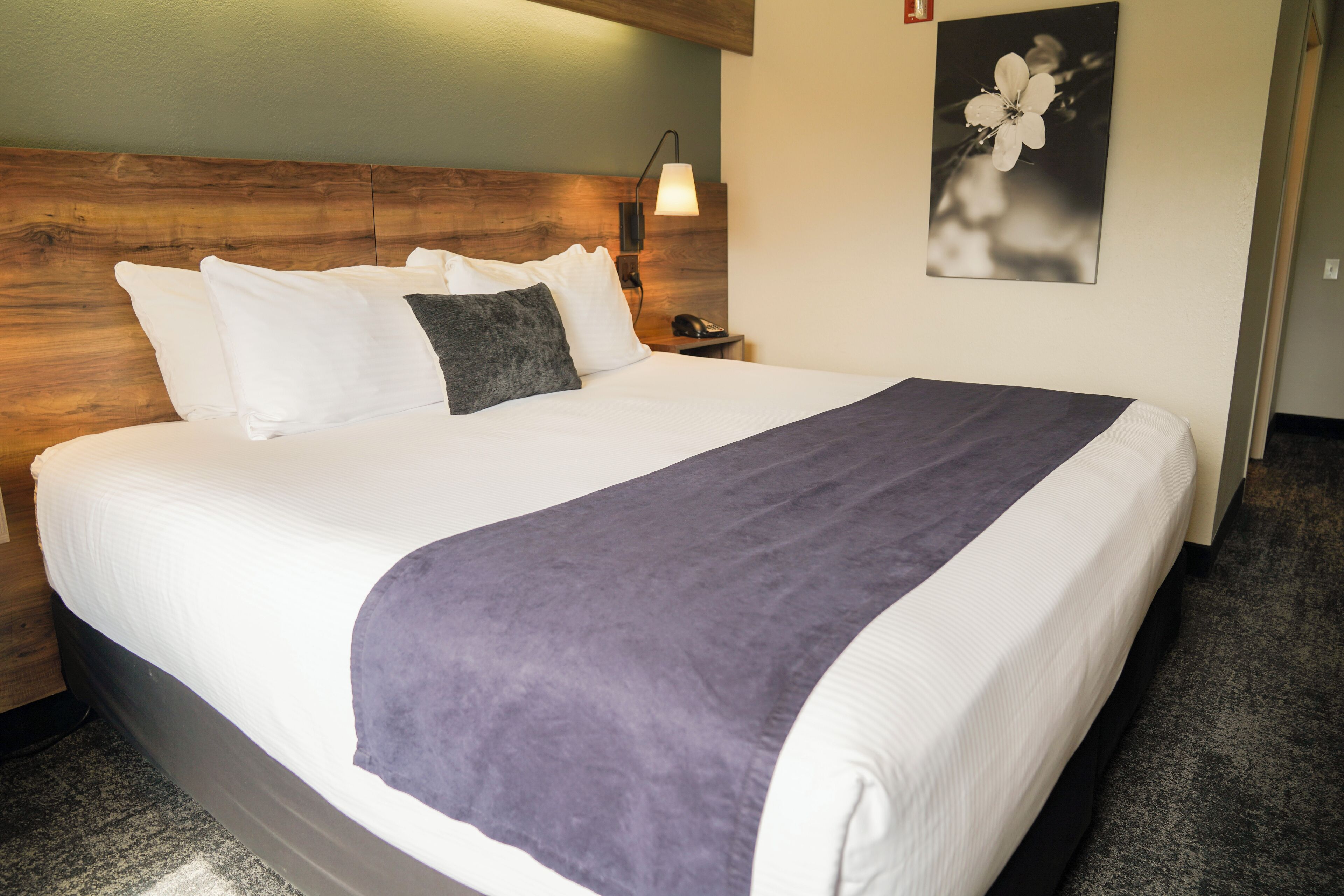 A neatly made bed with white and gray bedding, a wooden headboard, a bedside lamp, and a framed picture on the wall.