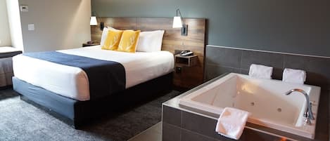 Ein modernes Hotelzimmer mit einem großen Bett, einem separaten Badezimmer mit Badewanne und einem kleinen Nachttischchen mit Lampe.