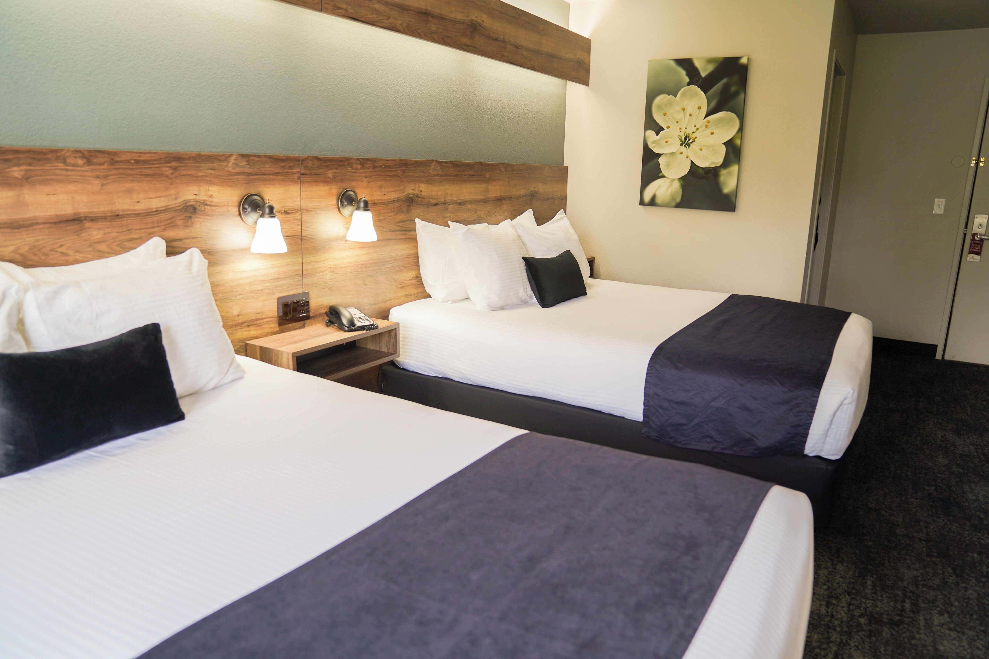 A hotel room with two beds, wooden headboard, bedside lamps, a painting of a flower, and a door.