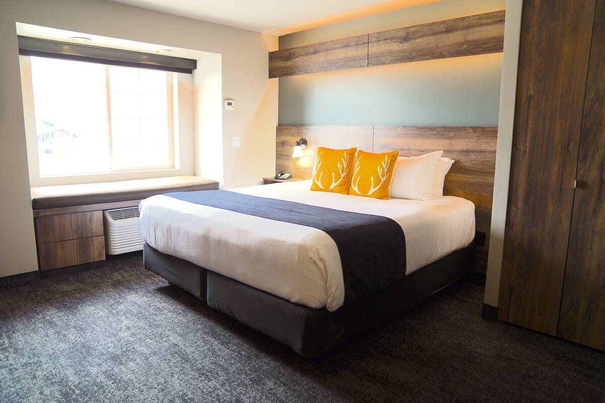 A modern hotel room with a large bed, a window with blinds, a bedside table with a lamp, and a wardrobe.