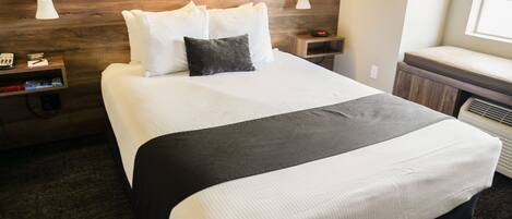 A hotel room with a large bed, wooden headboard, two bedside tables with lamps, a nightstand, and a window with blinds.
