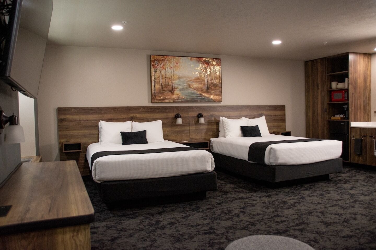 A hotel room with two beds, a painting on the wall, a wooden headboard, a desk, and a cabinet with a microwave.