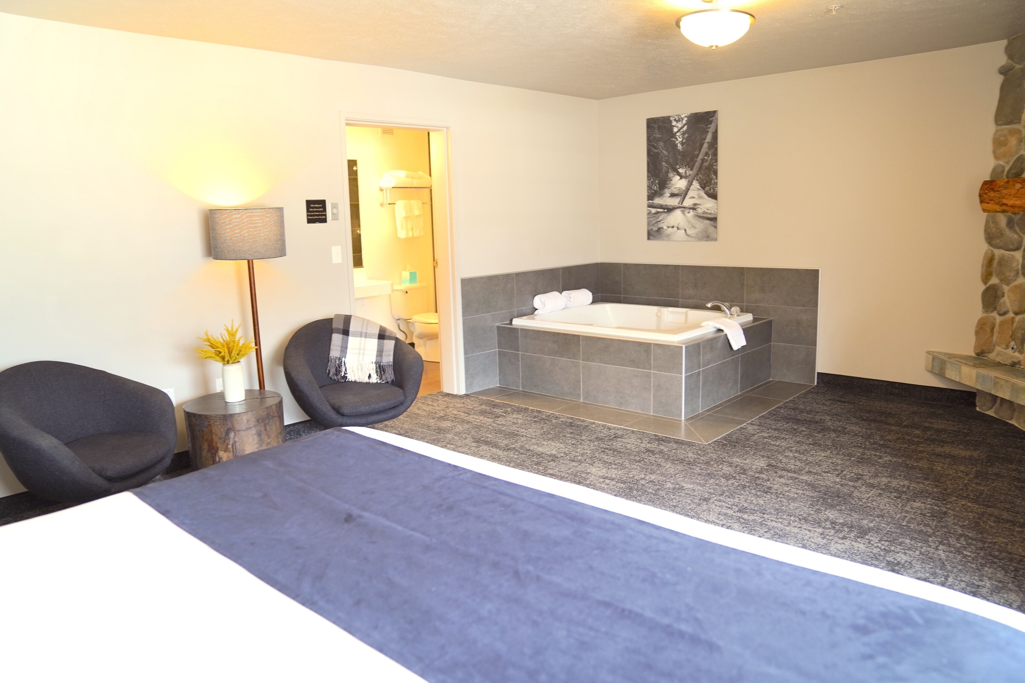 A modern hotel room with a large bed, a sitting area with a chair and a small table, a bathtub, and a stone wall.