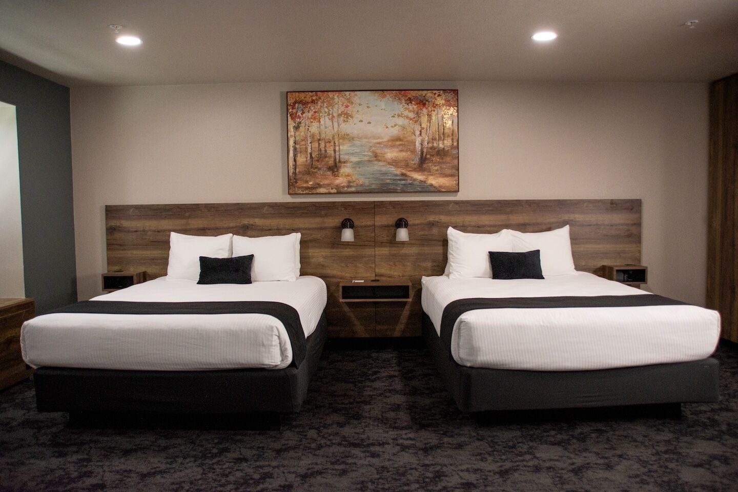 Quarto de hotel com duas camas, um quadro de uma paisagem florestal e cabeceira de madeira.