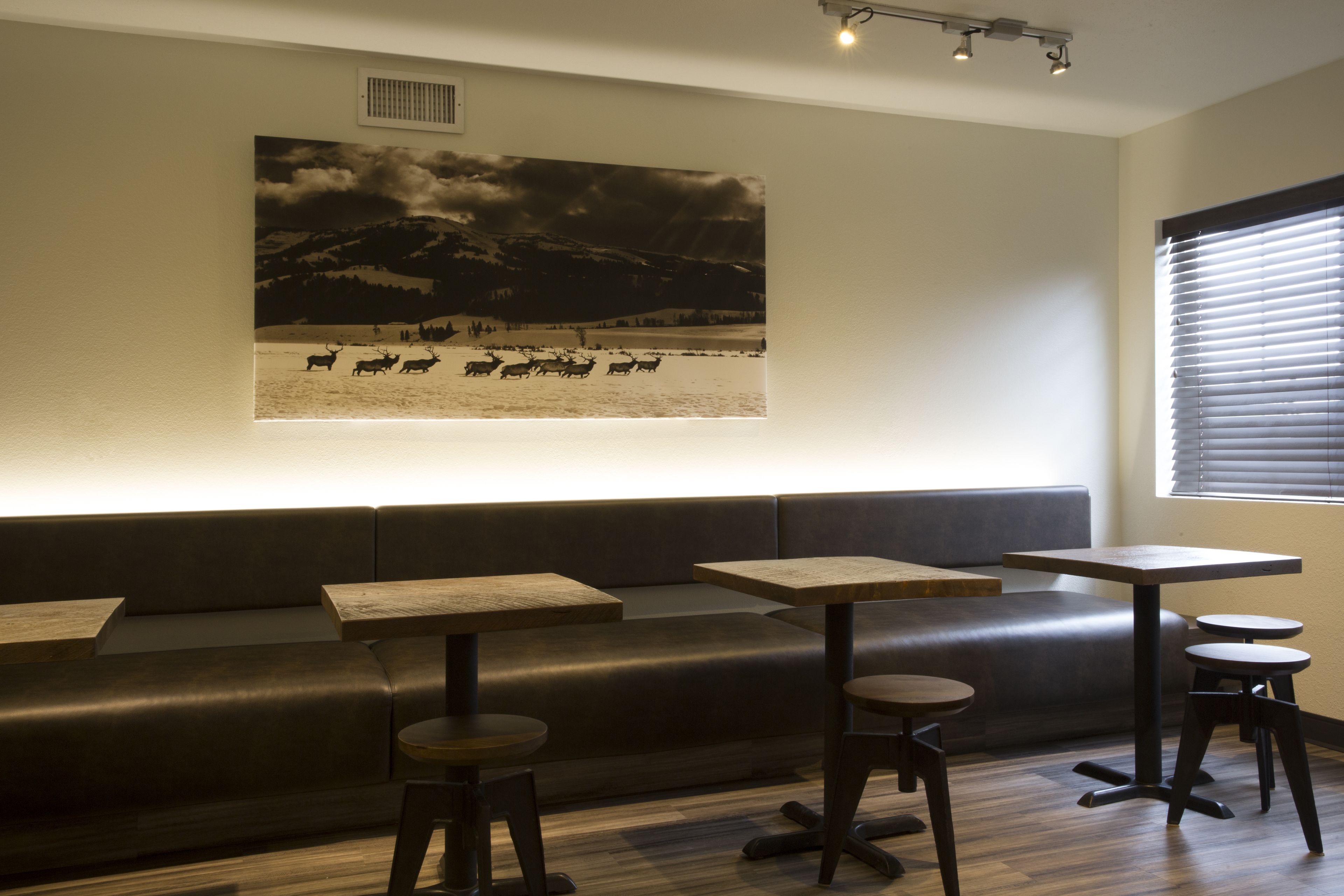 A modern interior with a dark leather sofa, wooden tables, and stools. A large black and white photograph of a snowy landscape with animals is displayed on the wall.