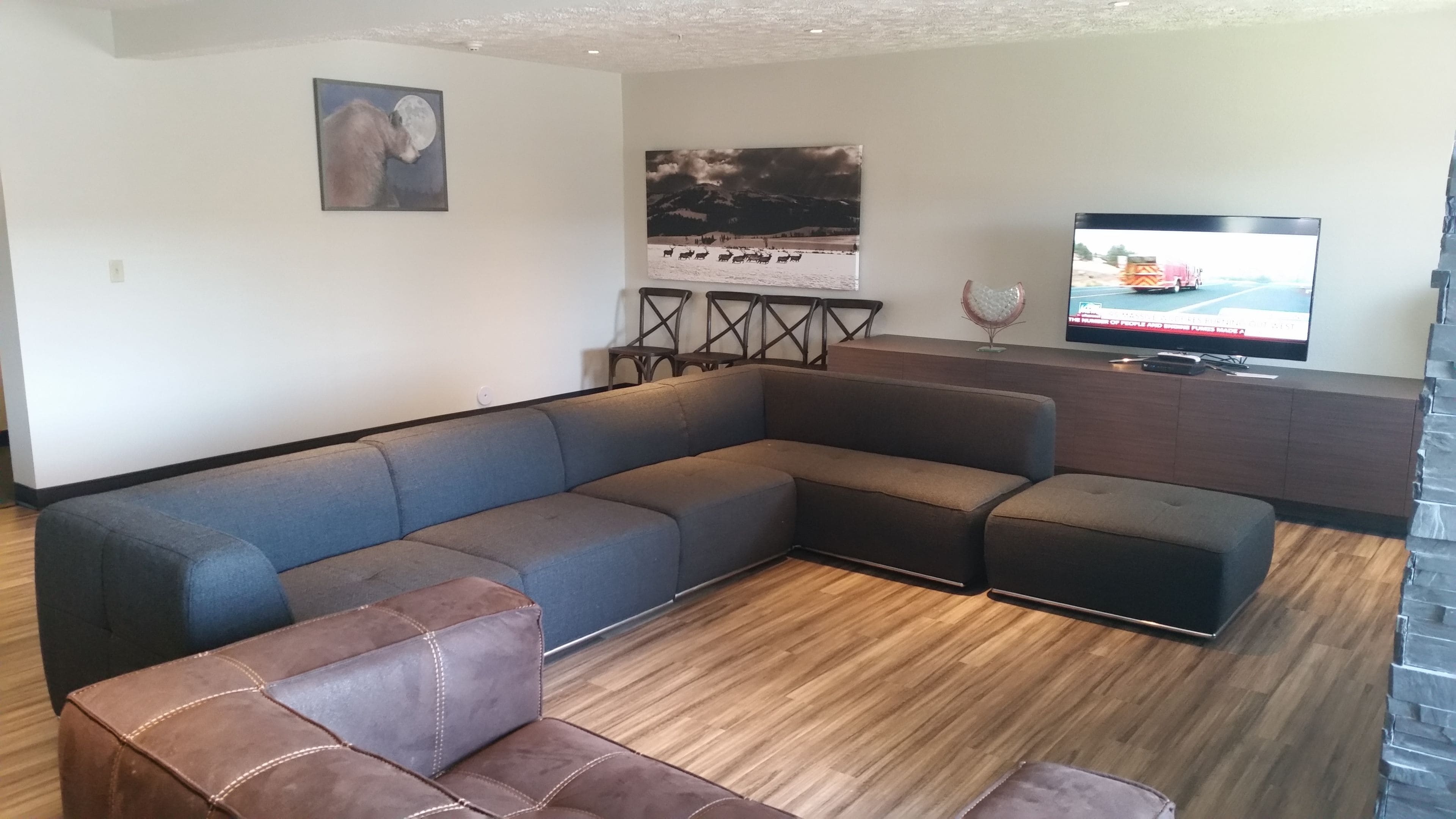 A modern living room with a sectional sofa, a small ottoman, a flat-screen TV, and a wall-mounted TV.