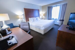 Et hotelværelse med seng, skrivebord med computer, stol, fjernsyn og et vindue med gardiner.