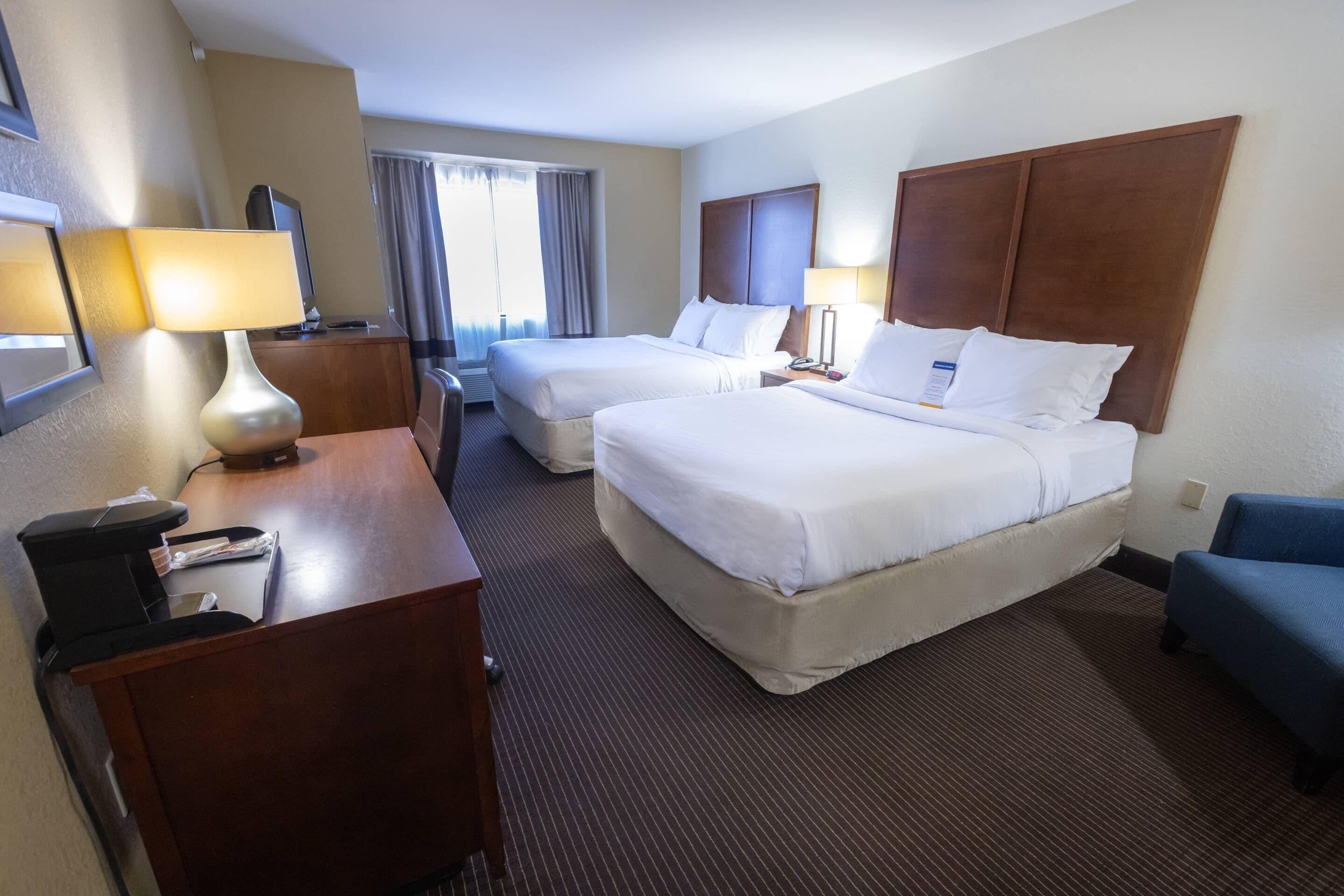 A hotel room with two beds, a desk with a lamp, a chair, and a window with curtains.