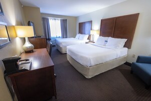Una habitación de hotel con dos camas, un escritorio con lámpara, una silla y un ventanal con cortinas.