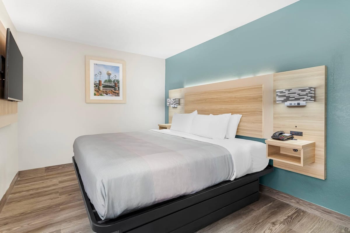A modern hotel room with a bed, a mounted TV, a framed picture on the wall, and a nightstand with a phone.