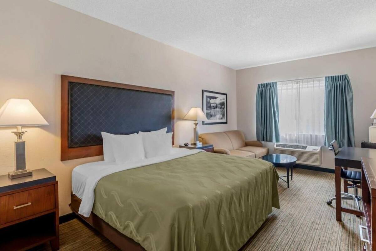 A hotel room with a bed, bedside lamps, a desk, a chair, a sofa, and a window with curtains.