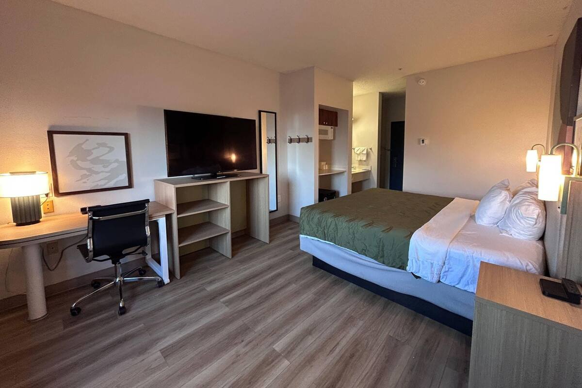 A hotel room with a bed, a desk with a chair, a TV, and a bathroom visible through an open door.
