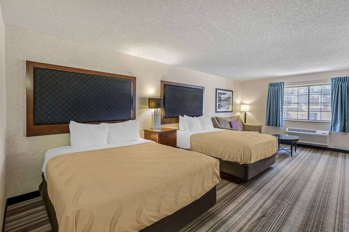 A hotel room with two beds, a desk, a chair, a TV, a window with curtains, and an air conditioning unit.